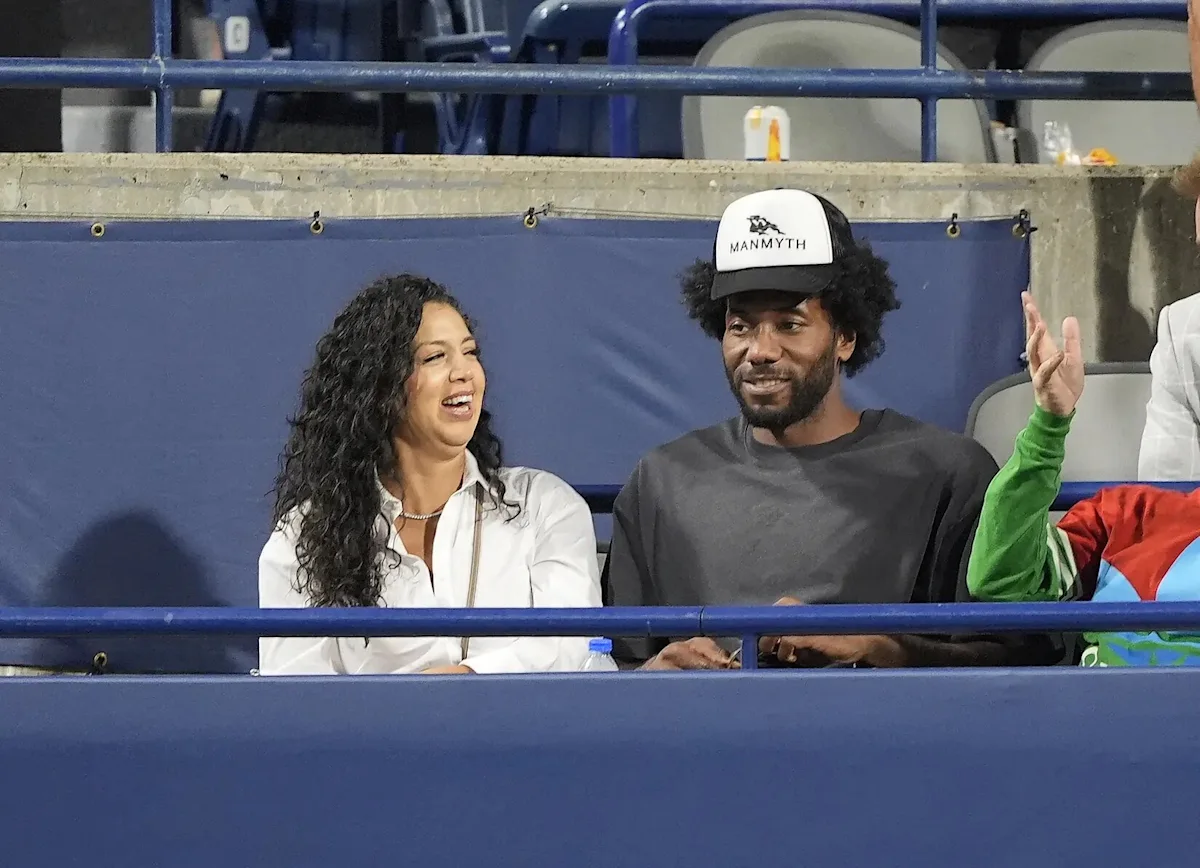 Woman laughing beside a bearded man wearing a cap in arena seats behind blue railings