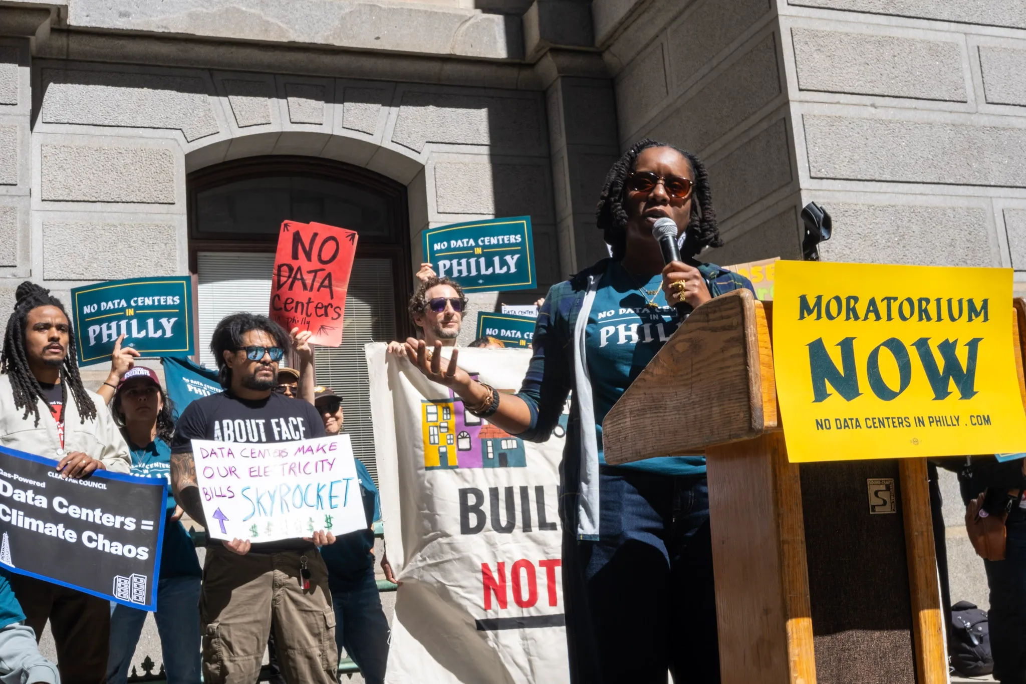 Protesters hold anti-data-center signs as a speaker addresses a crowd outside a stone building.