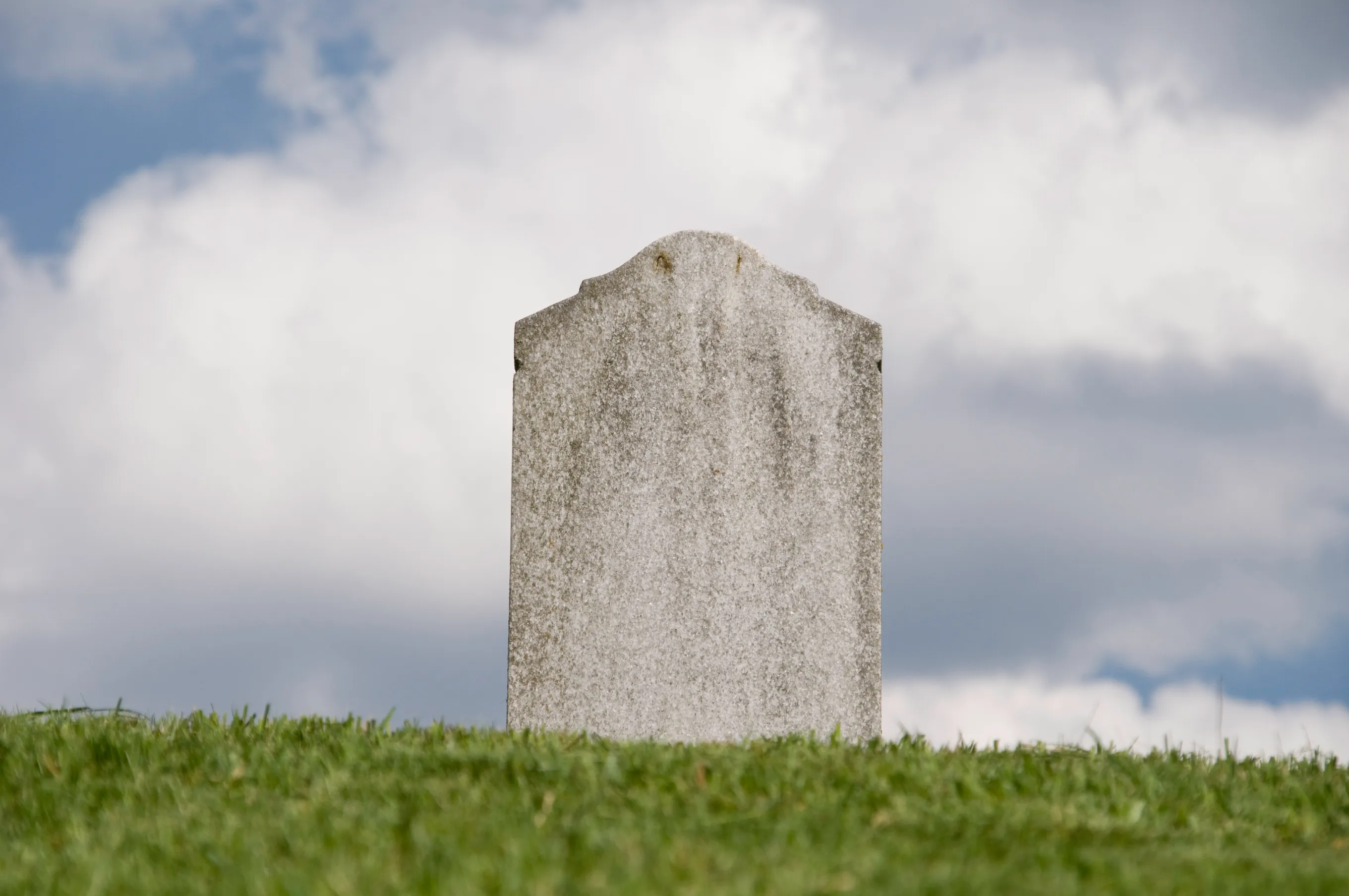 Weathered gravestone standing in a grassy field beneath a cloudy blue sky