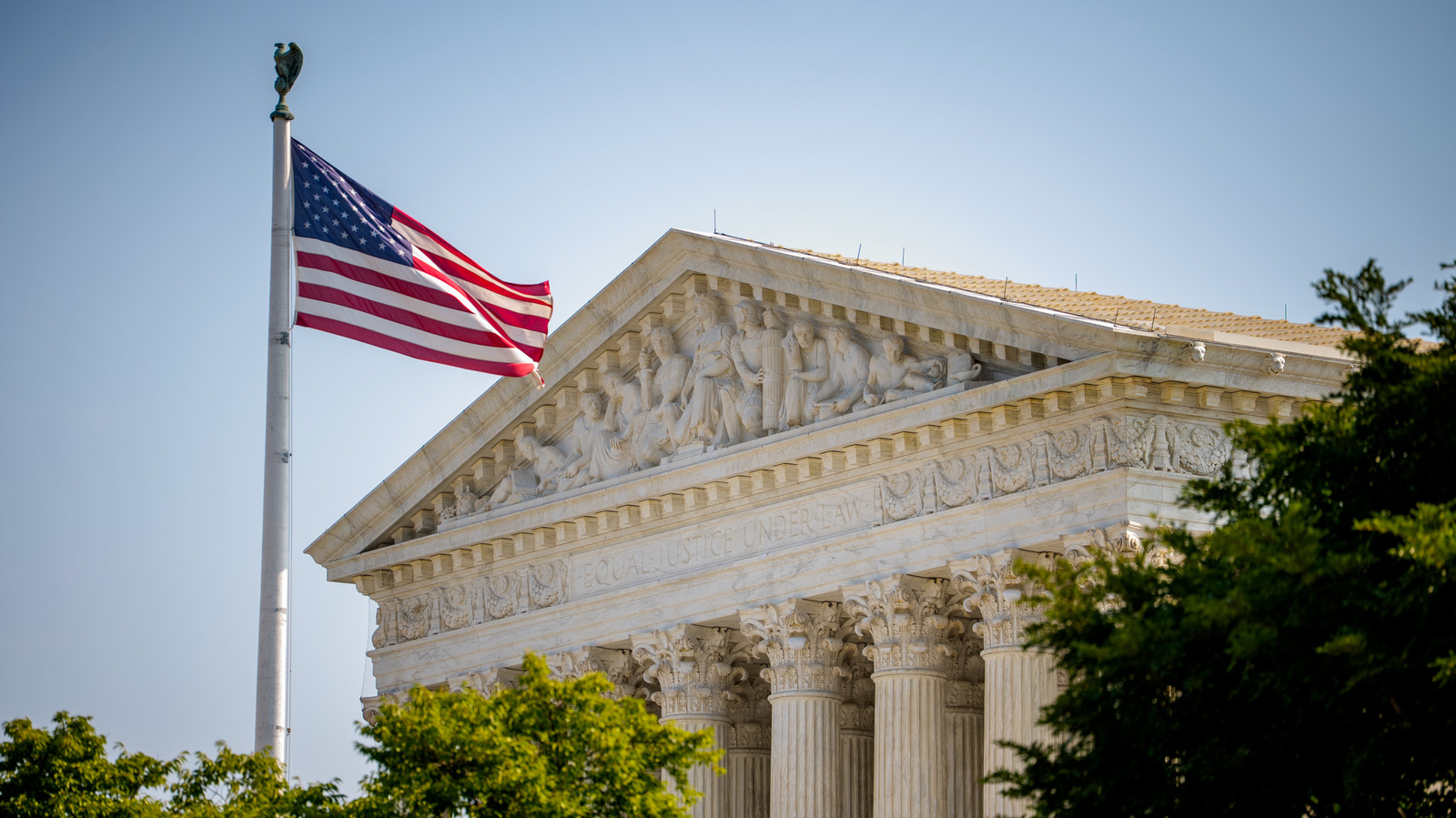 An American flag waves beside the sculpted pediment and columns of a courthouse in daylight.