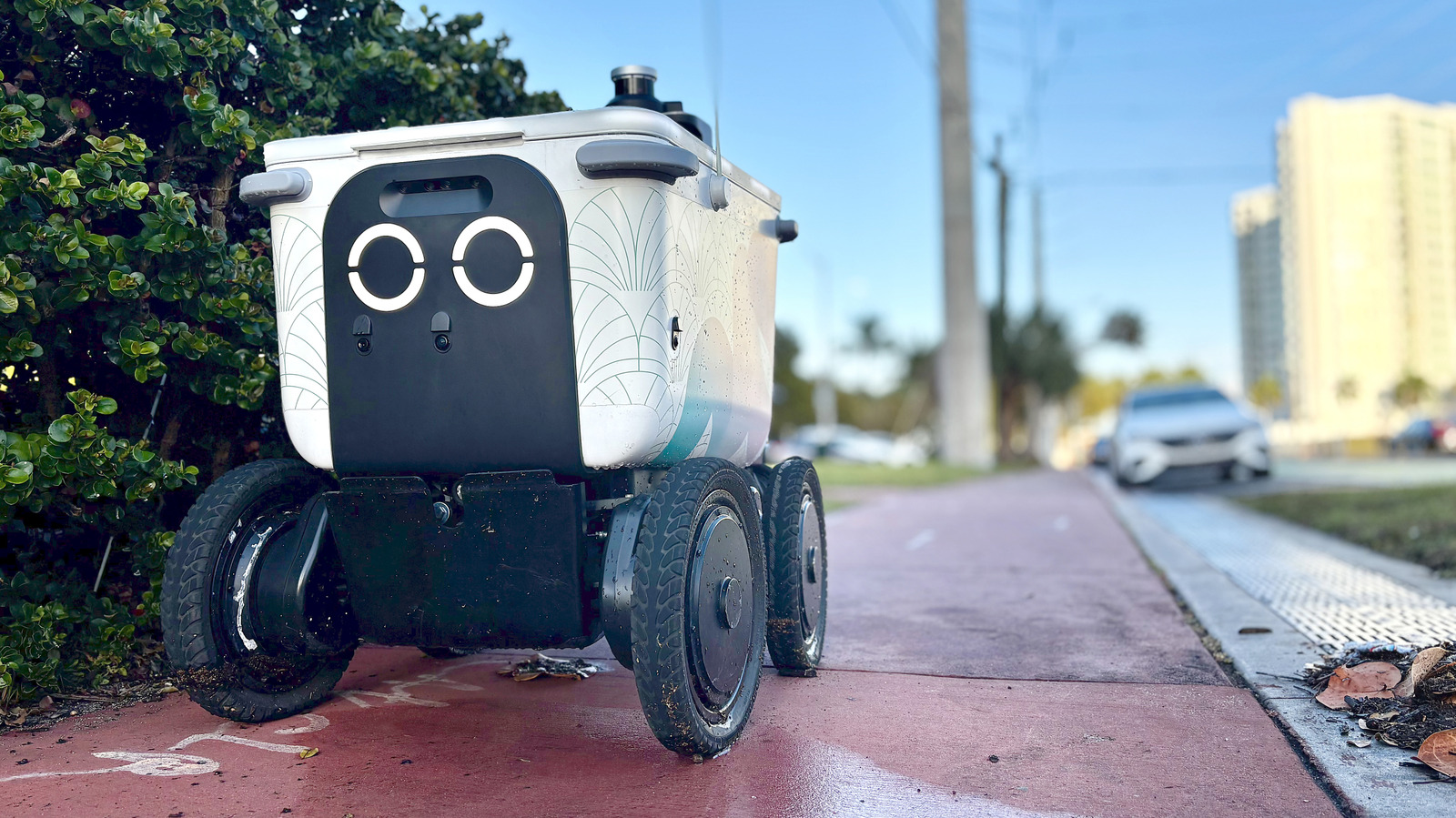 A small four-wheeled delivery robot sits on a wet city sidewalk beside greenery.