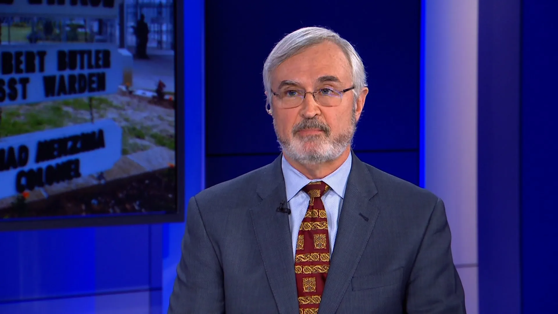 Gray-haired man in glasses and suit seated in a blue-lit television studio.