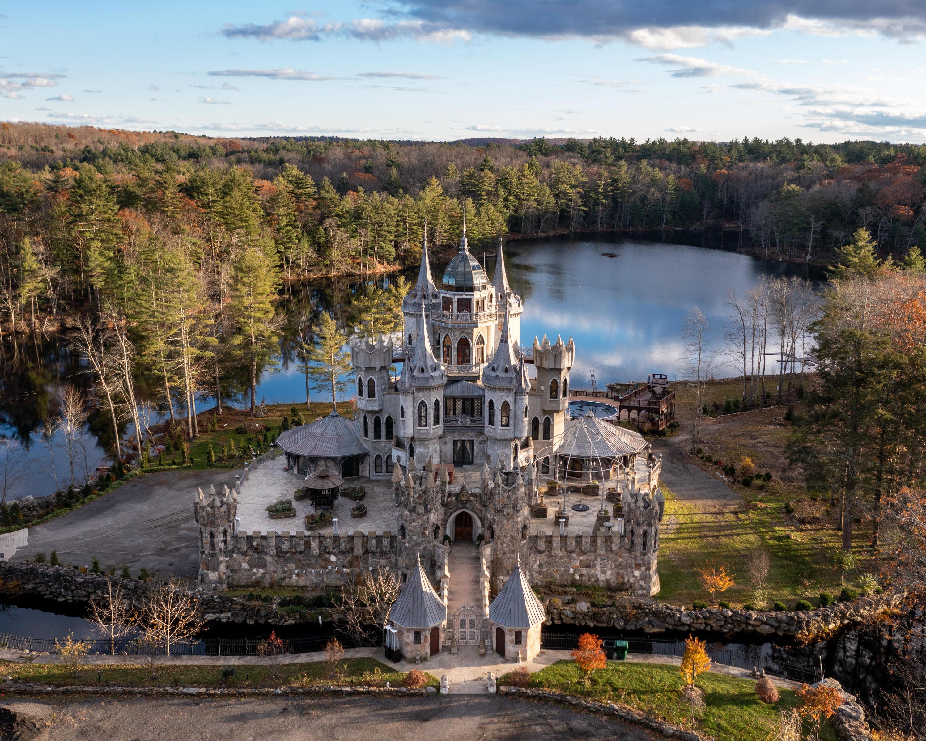 A stone castle stands beside a wooded lake beneath a partly cloudy sky.