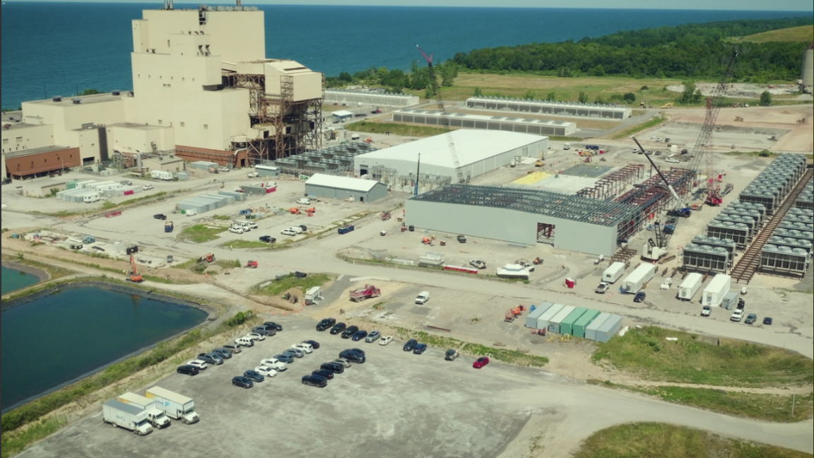 Aerial view of a large industrial facility under construction beside a body of water, with cranes and parked vehicles.