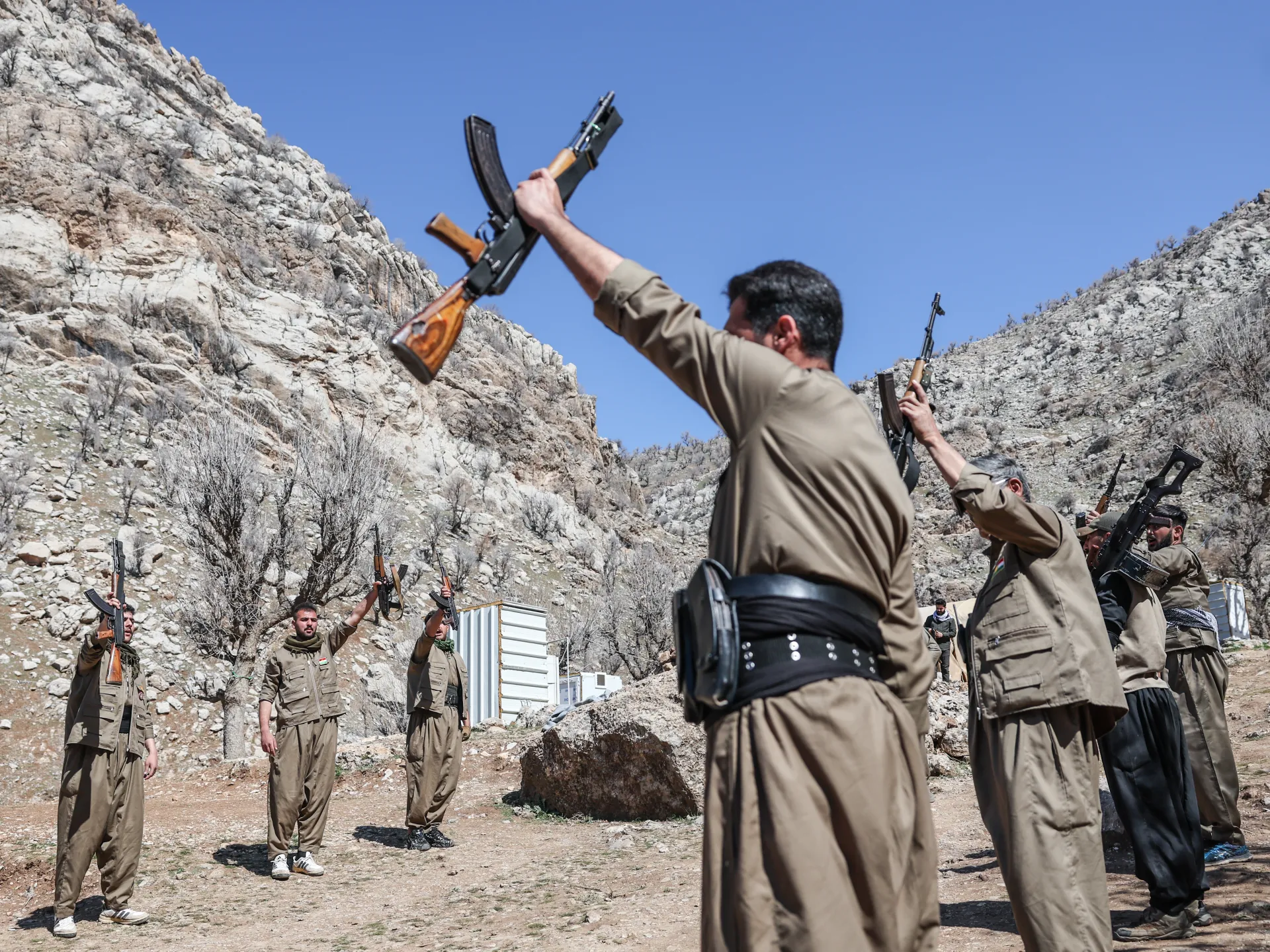 Armed men in tan uniforms raise rifles in a rocky mountain valley beneath a clear blue sky.