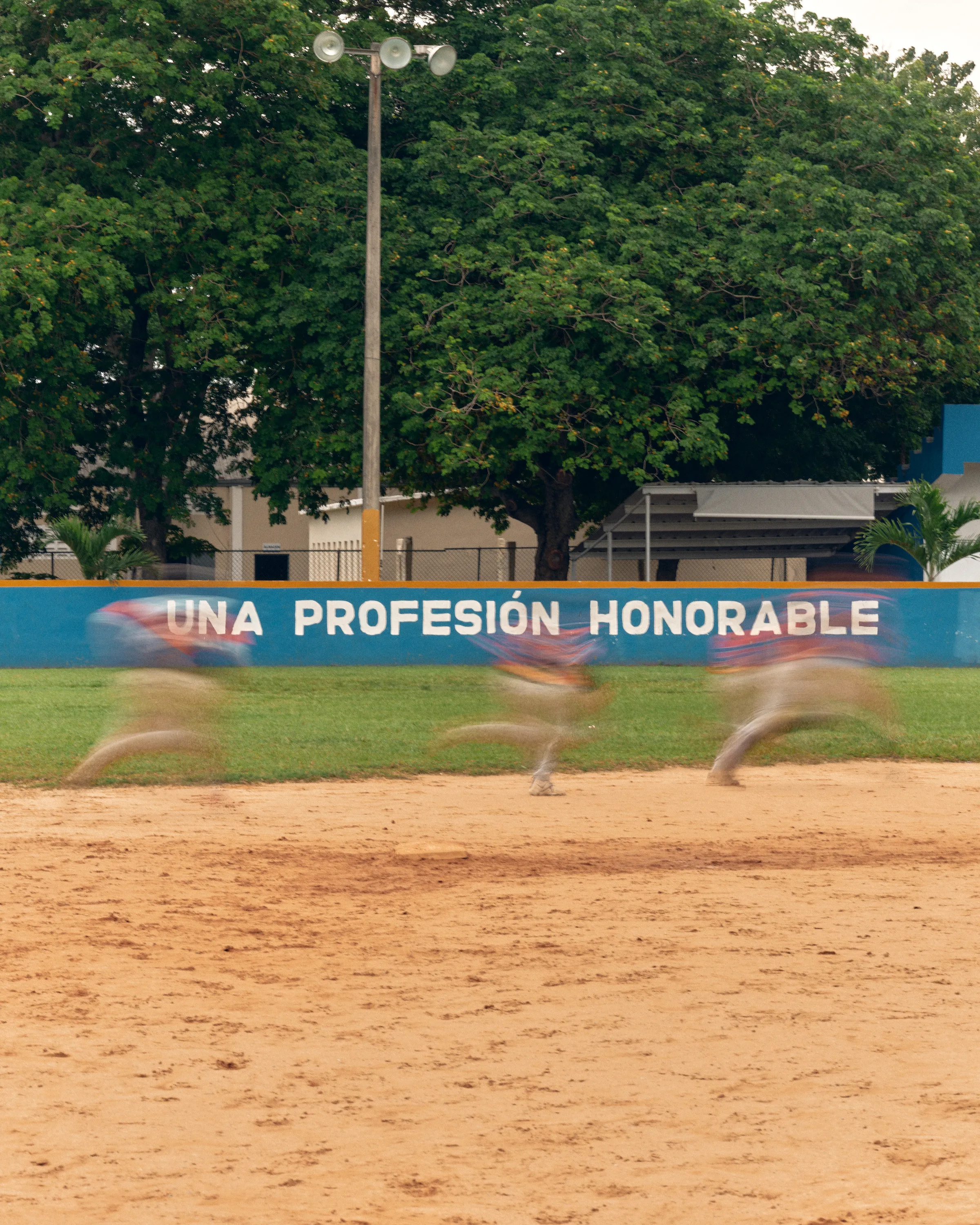 Blurred baseball players run past a blue wall reading “UNA PROFESIÓN HONORABLE” on a field.