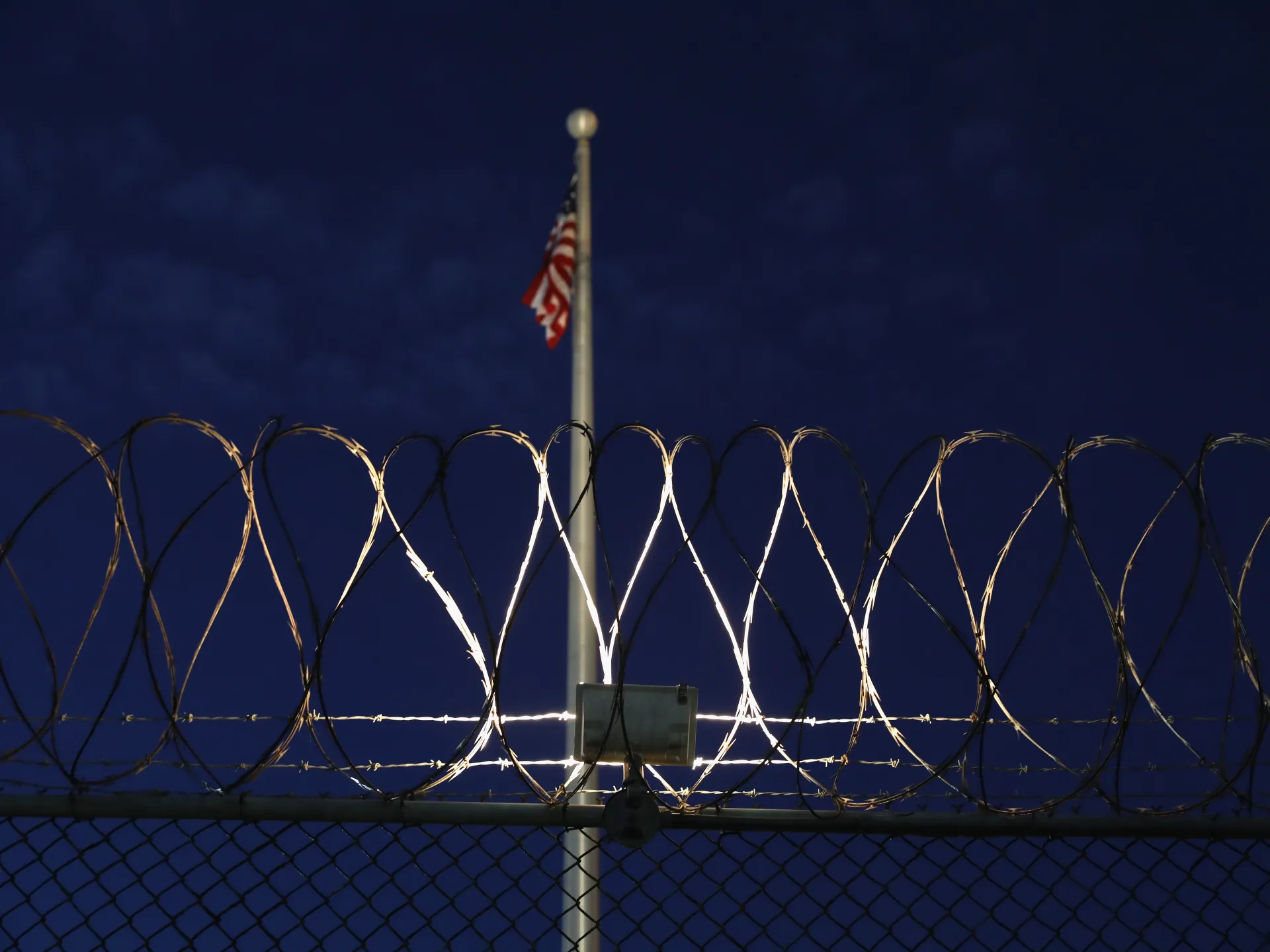 American flag behind razor wire and chain-link fencing against a dark blue night sky