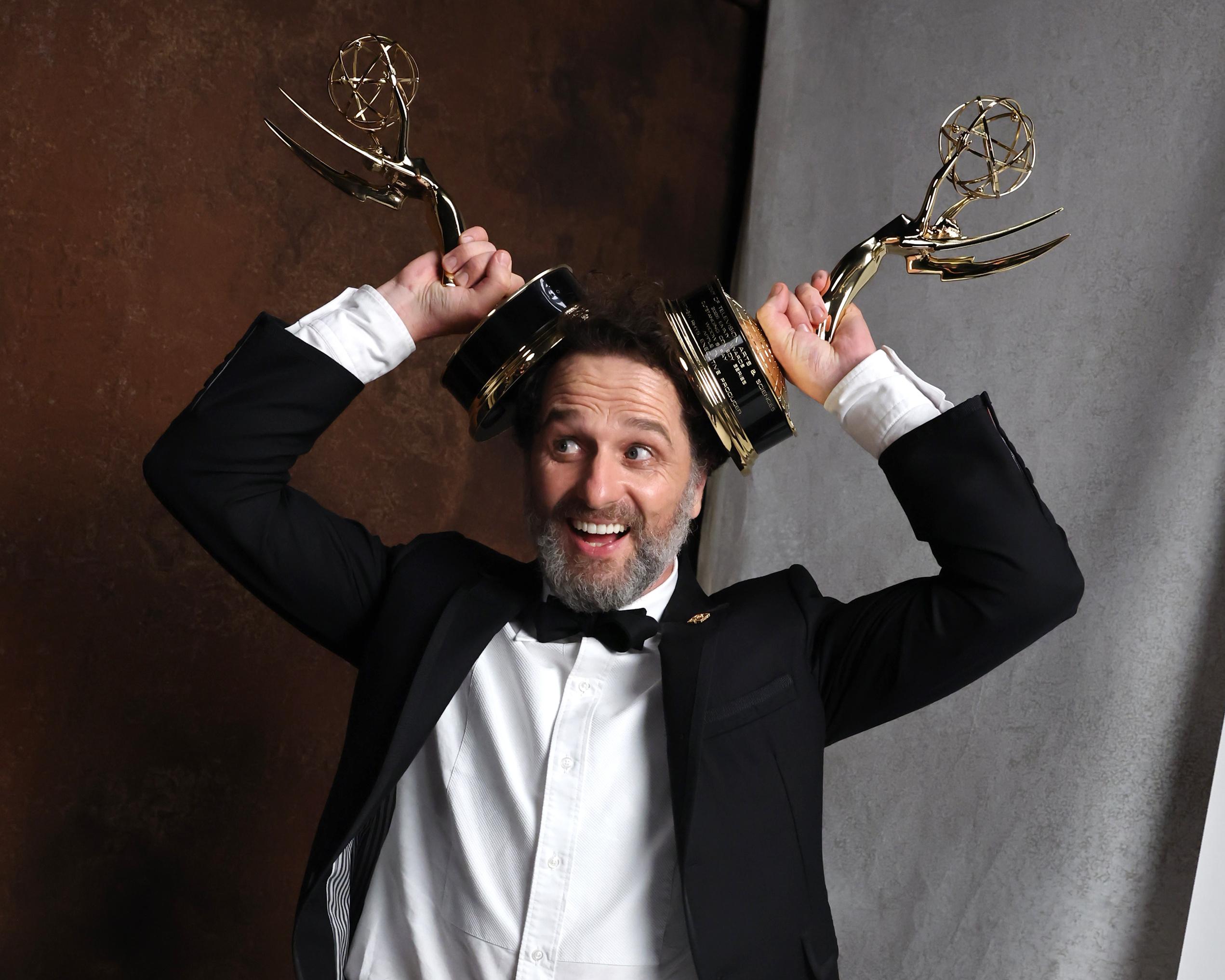 Smiling man in tuxedo raises two Emmy statuettes above his head.