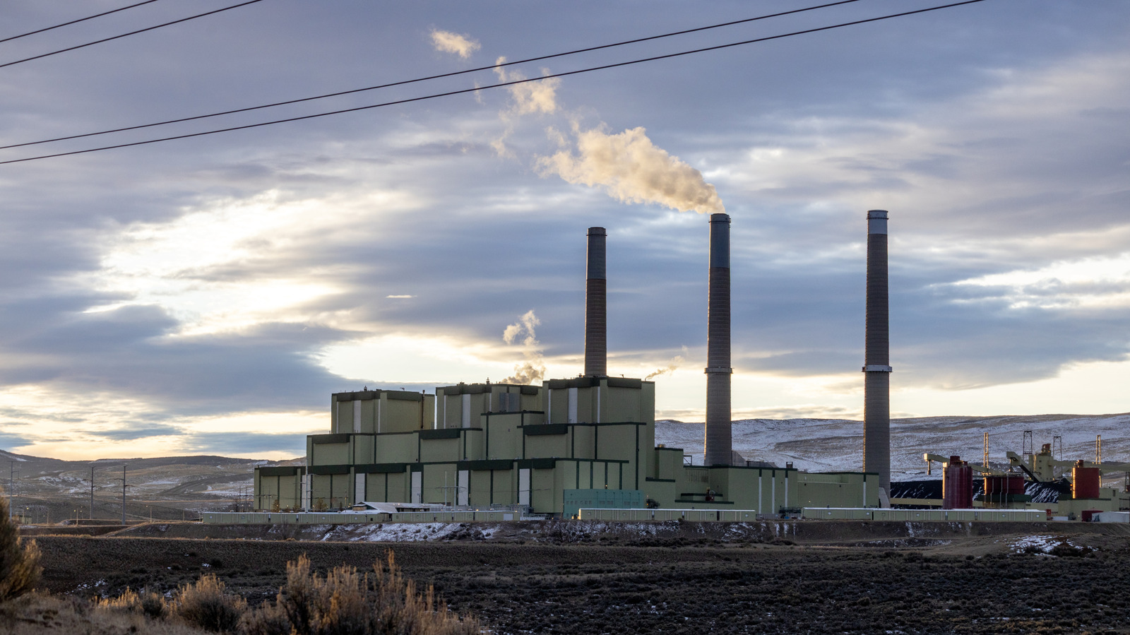 Power plant with three smokestacks emitting steam against a snowy, mountainous landscape