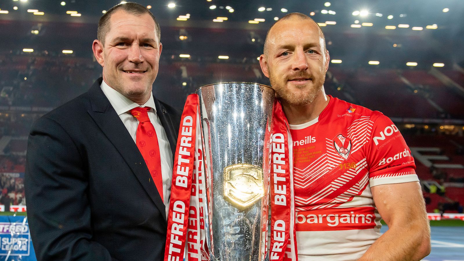 Two men pose with a large silver rugby trophy inside a stadium.