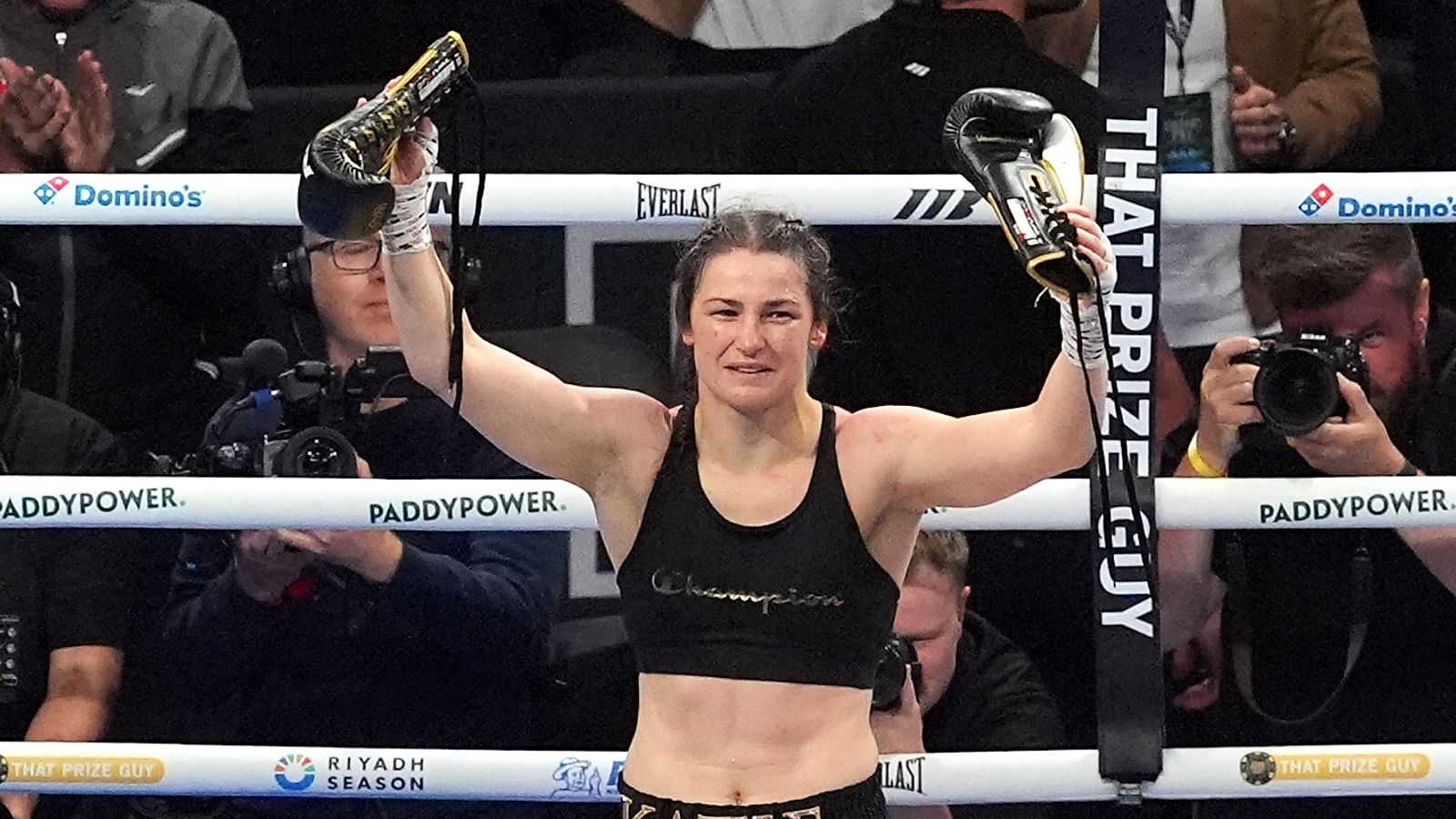Female boxer raises both gloved fists inside a boxing ring as photographers capture the moment.