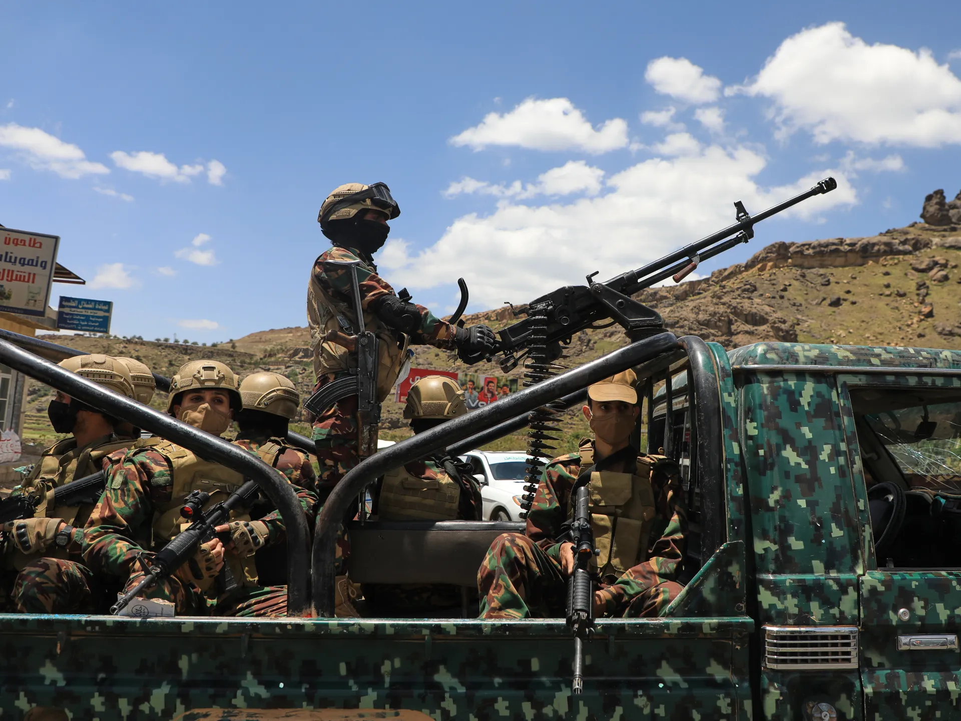 Armed soldiers ride in a camouflage-painted pickup truck beneath a bright, cloud-filled sky.