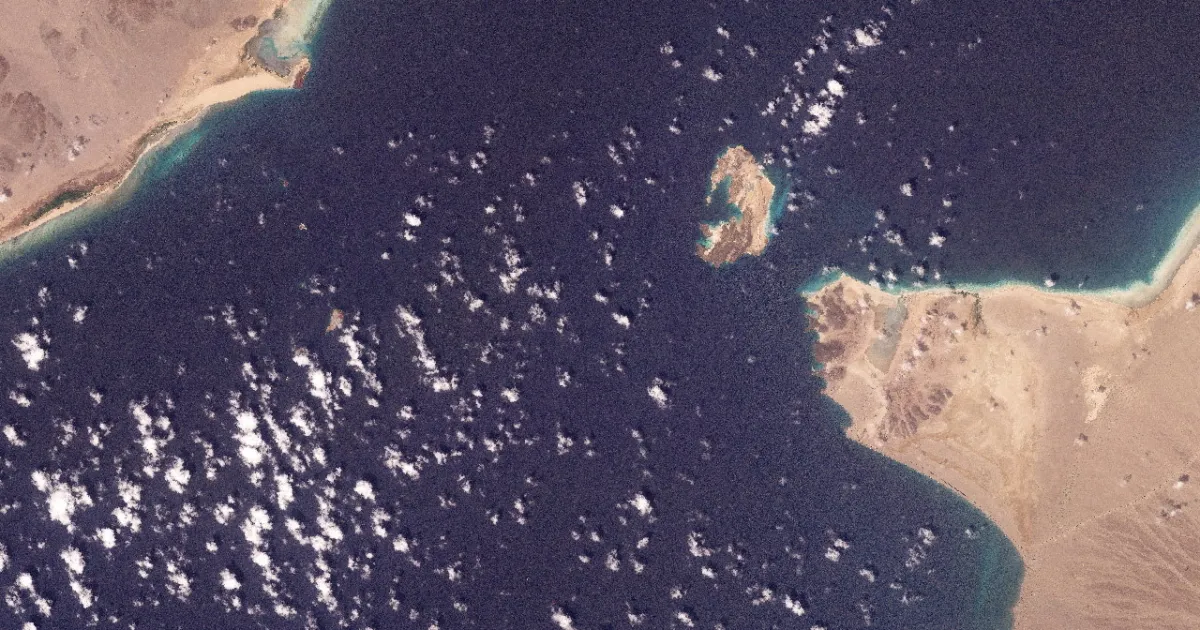 Satellite view of a small rocky island between desert coastlines, surrounded by dark blue water and scattered clouds.
