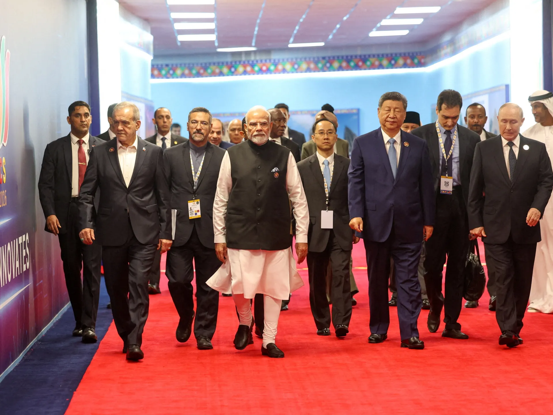 Group of suited officials walking together along a red carpet inside a brightly decorated conference venue