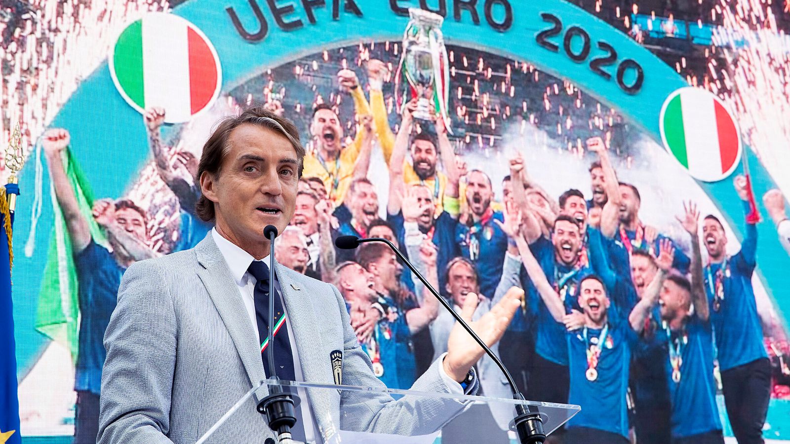 A suited man speaks at a lectern before cheering football supporters in blue shirts and Italian flags.