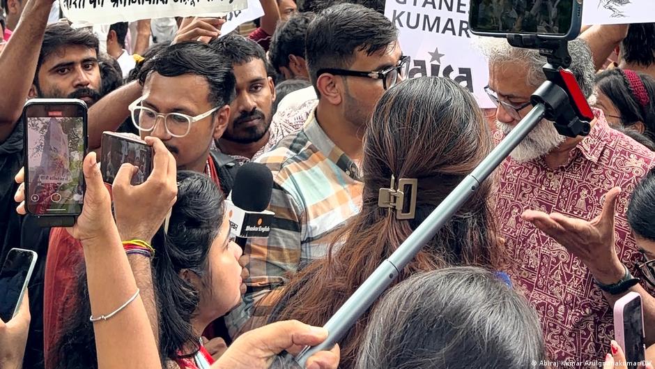 Crowd surrounds an older man as people hold up phones and a microphone during a protest.