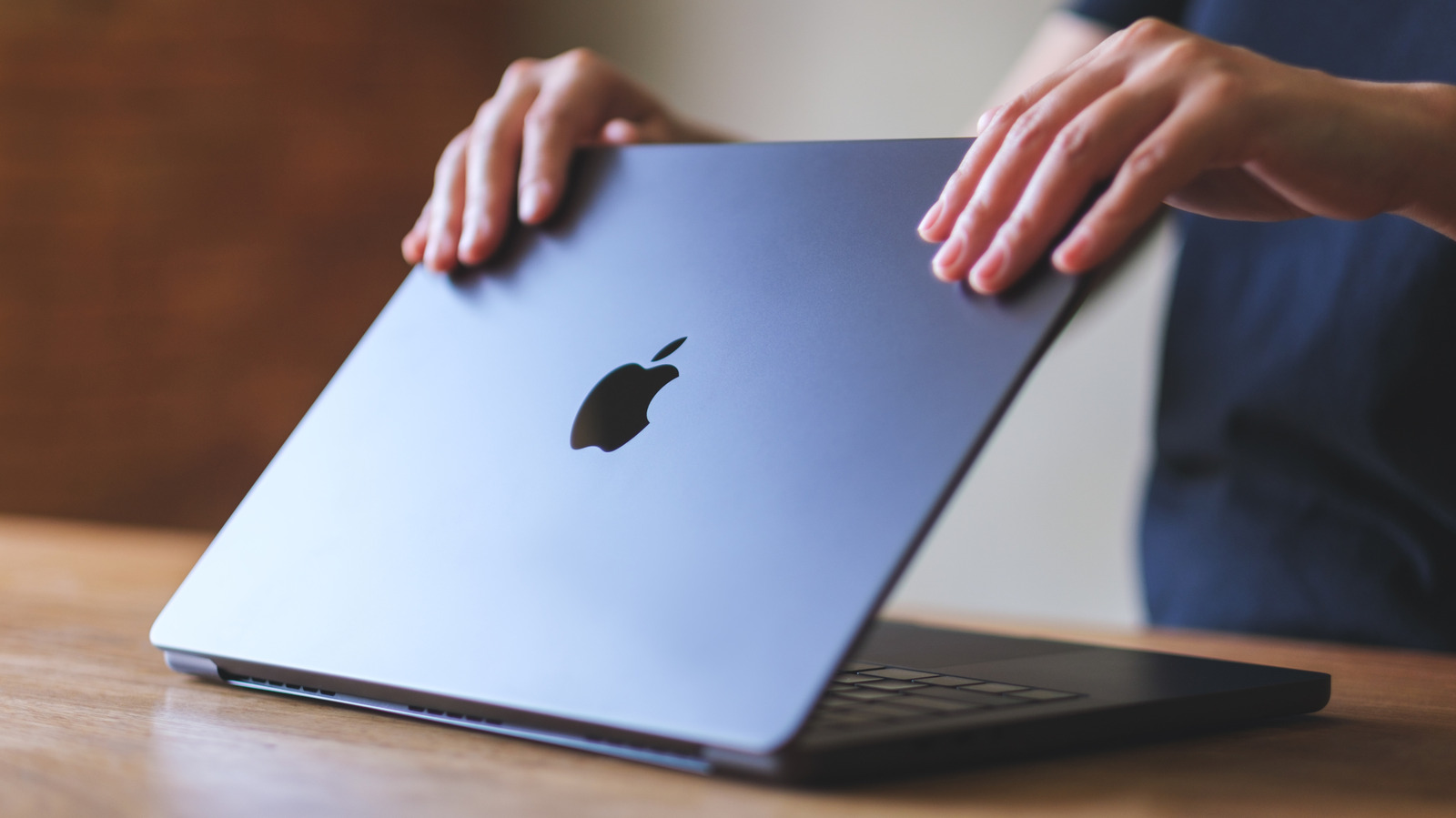 Hands lift the lid of a silver Apple laptop on a wooden table.