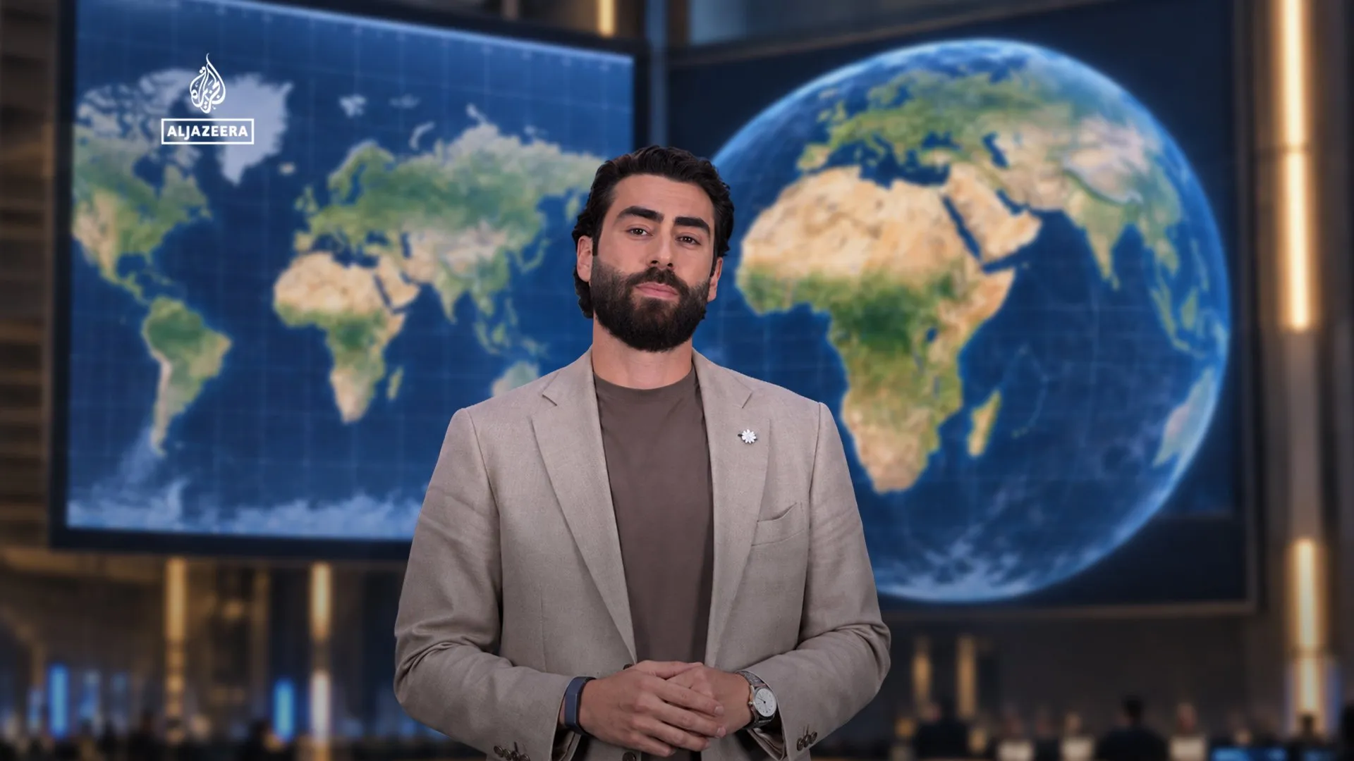 Bearded presenter in blazer stands before large digital world maps in a broadcast studio.