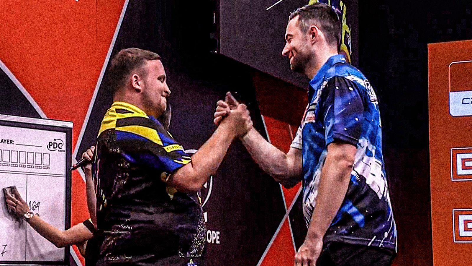 Two darts players in blue and yellow shirts shake hands on stage after a match.