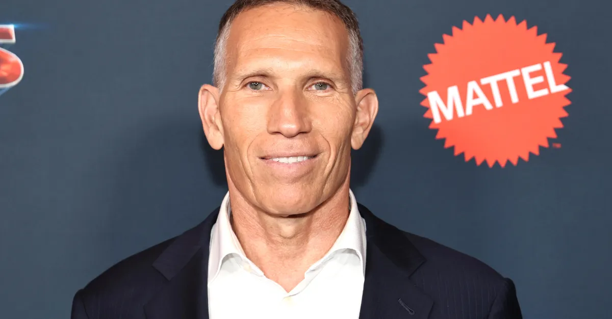 A man in a dark suit and white shirt poses before a Mattel logo backdrop.