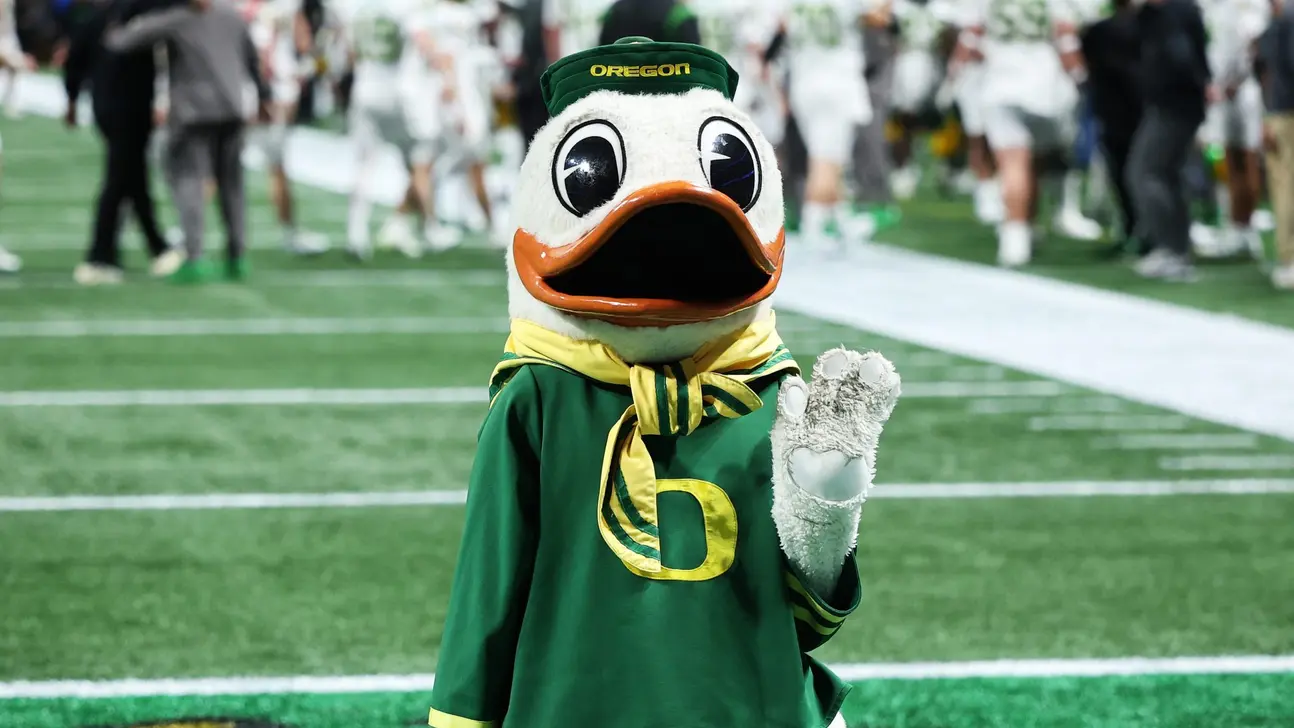 Oregon Duck mascot wearing a green uniform and hat, waving on a football field.