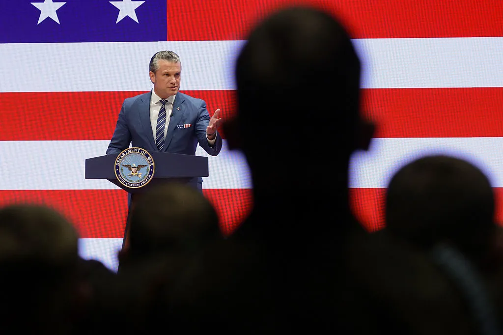 A suited man speaks at a Pentagon podium before a large U.S. flag, partly obscured by audience silhouettes.
