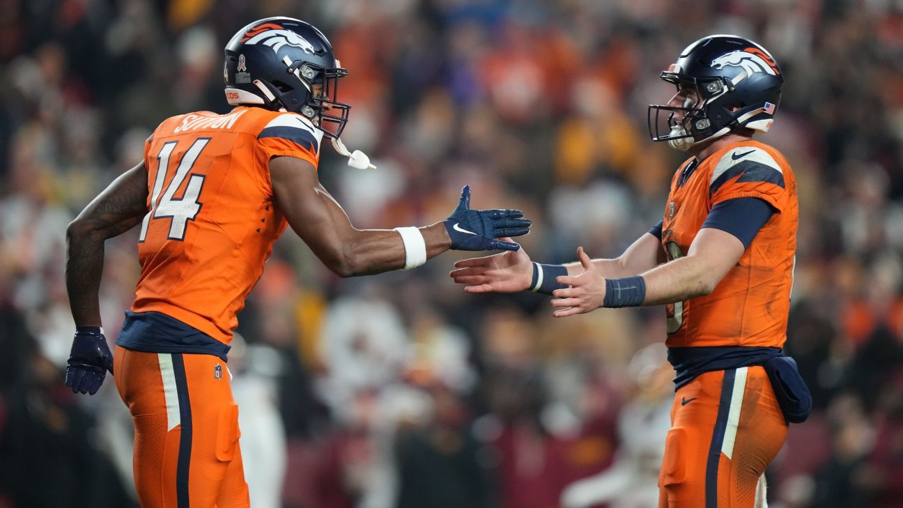 Two Denver Broncos players in orange uniforms reach toward each other on the field during a game.