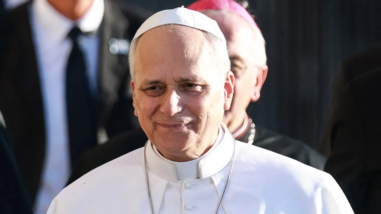 A man in white papal clothing smiles slightly, with clergy and other people behind him.
