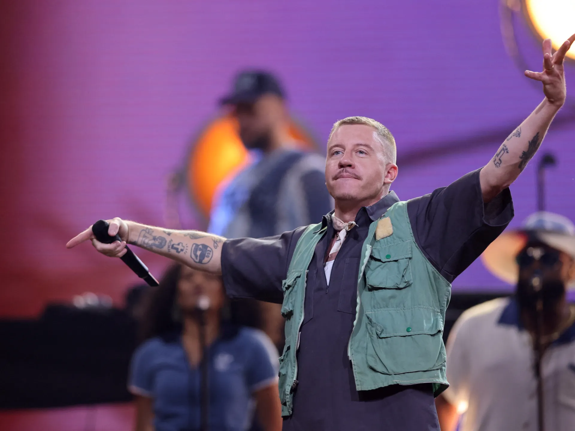 Tattooed male performer holds a microphone and raises both arms on a brightly lit stage.