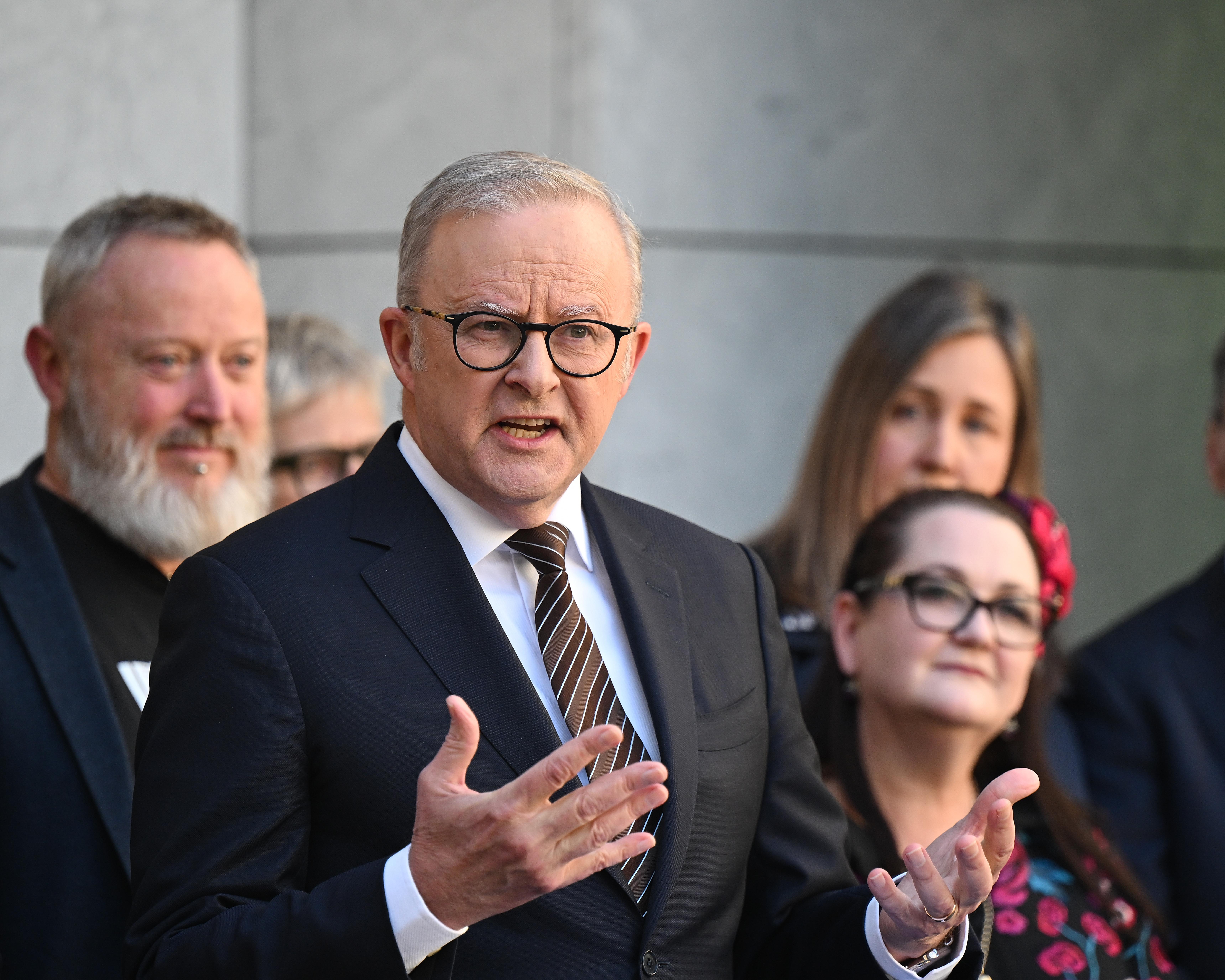 Grey-haired man in glasses and suit gestures while speaking among several people