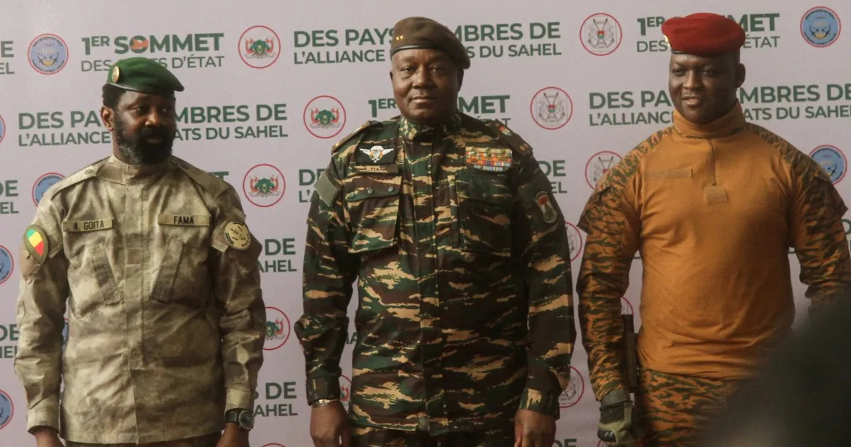 Three uniformed military officials stand before a backdrop promoting a Sahel states alliance summit.