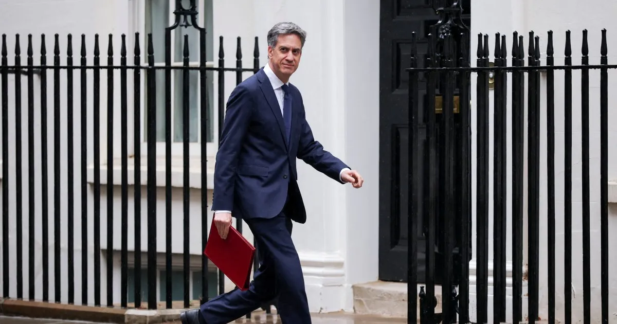 Man in navy suit carrying a red folder walks past black railings outside a building.