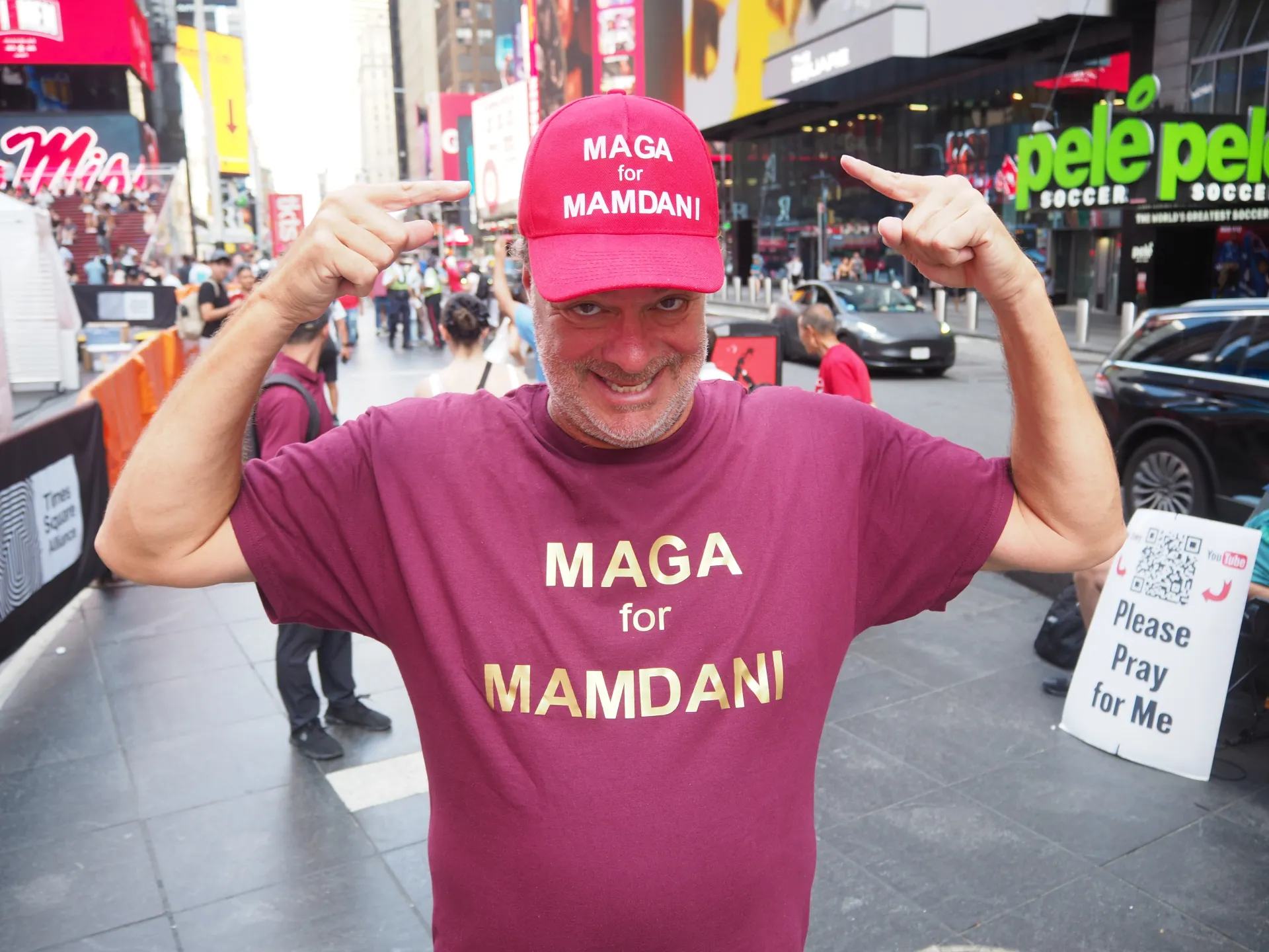 Man wearing a red MAGA for Mamdani cap and shirt, pointing at the messages in Times Square.