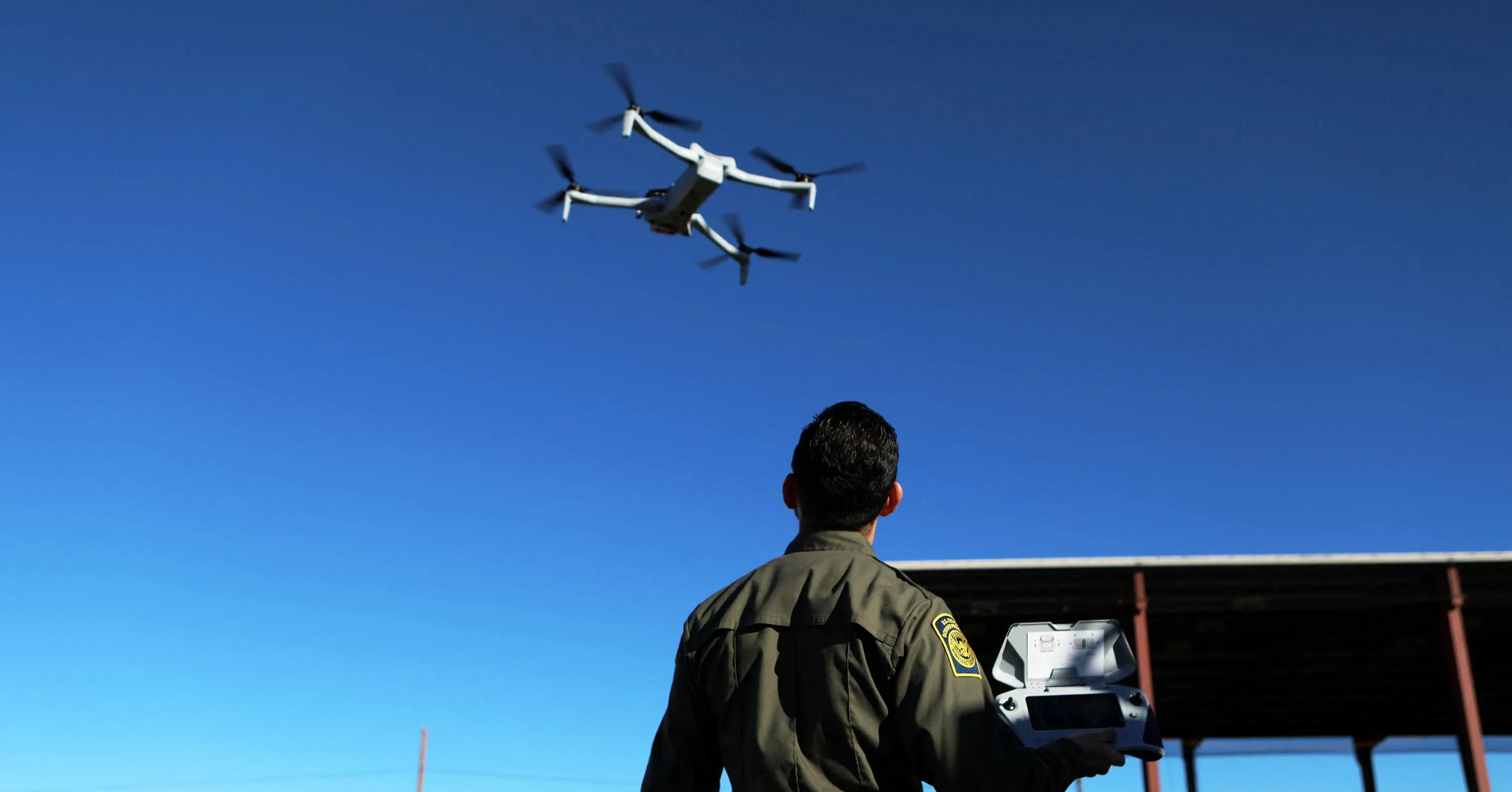 A uniformed man operates a handheld controller as a quadcopter flies overhead against a blue sky.