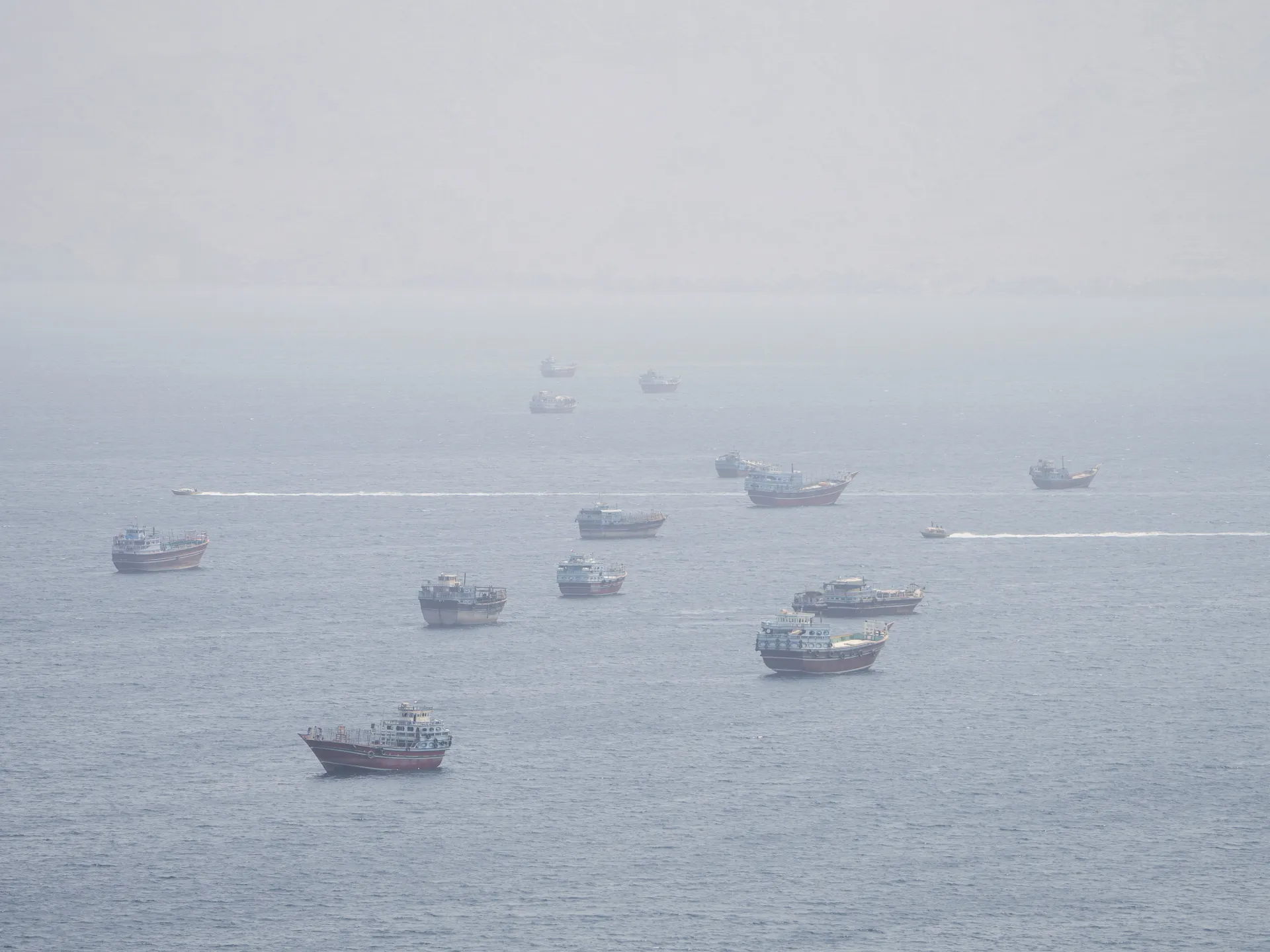 Several fishing boats and small vessels move across a hazy, calm sea.