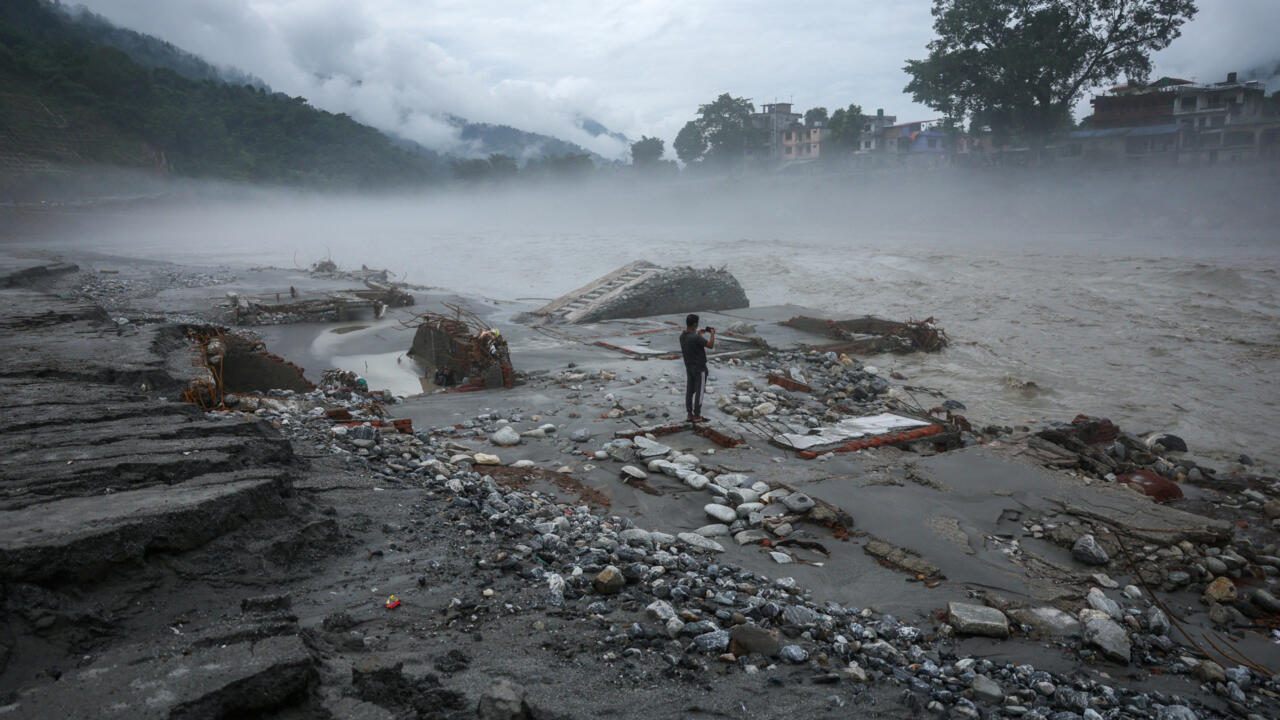 Floodwaters surround debris and damaged structures as a person photographs the scene beneath misty mountains.