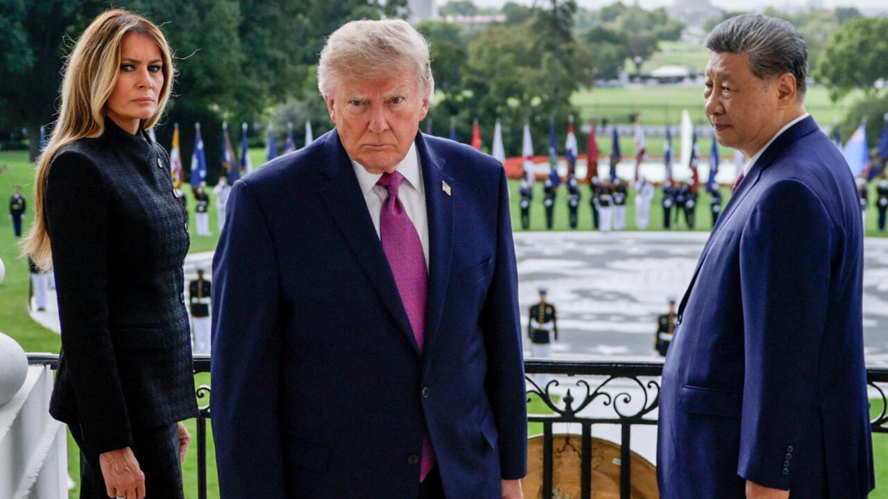 Three formally dressed people stand on a balcony overlooking a lawn lined with flags and ceremonial guards.
