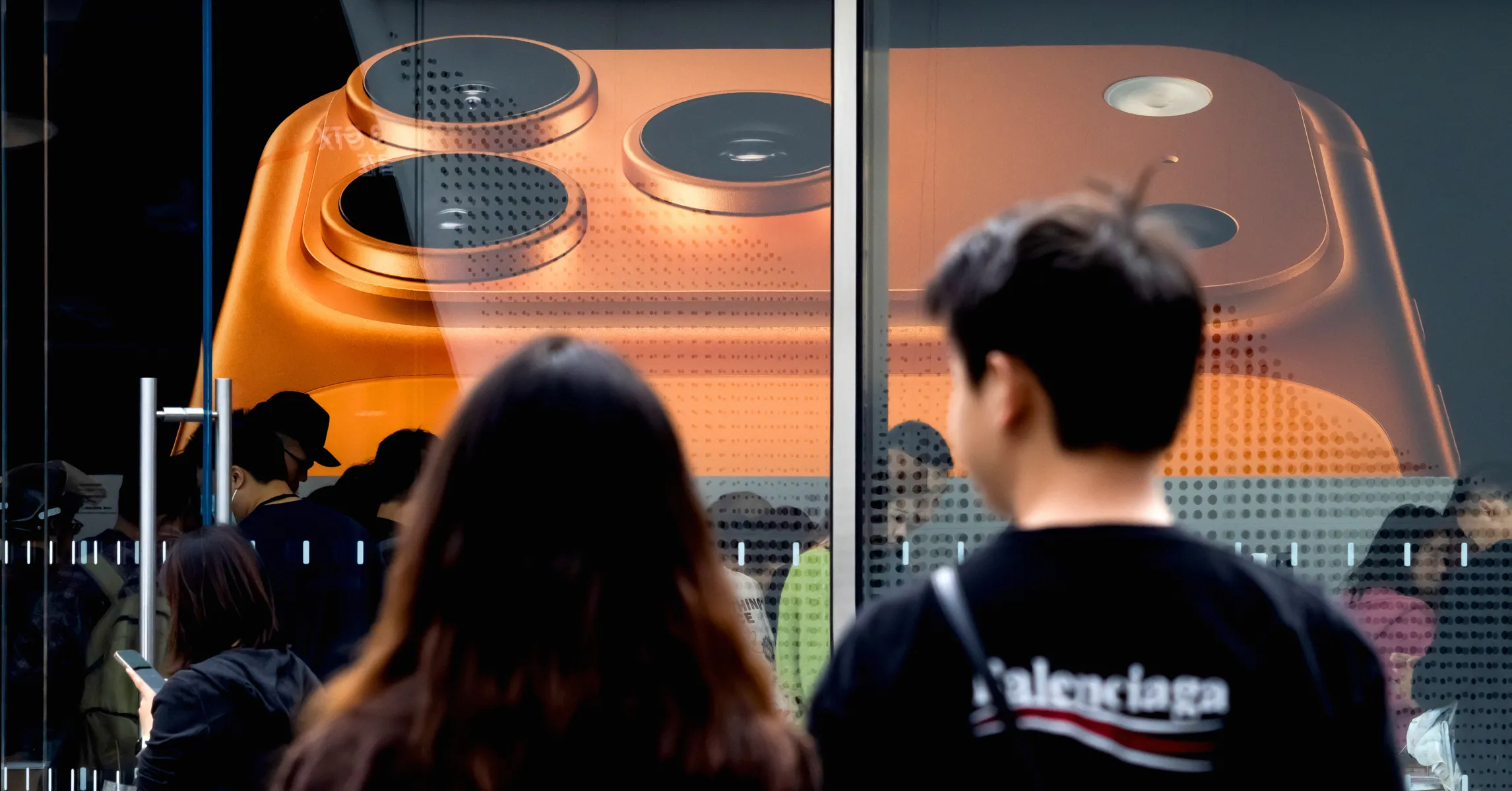 People view a large orange smartphone display through a storefront window.
