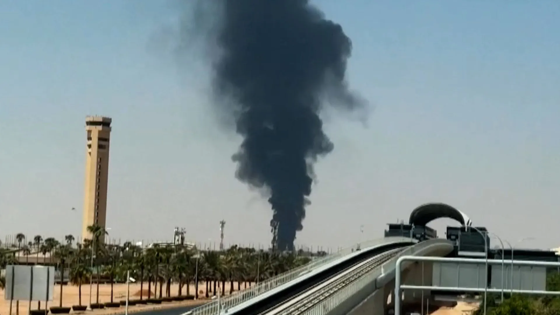Dark smoke plume rising behind palm trees beside an elevated transit track and tower.