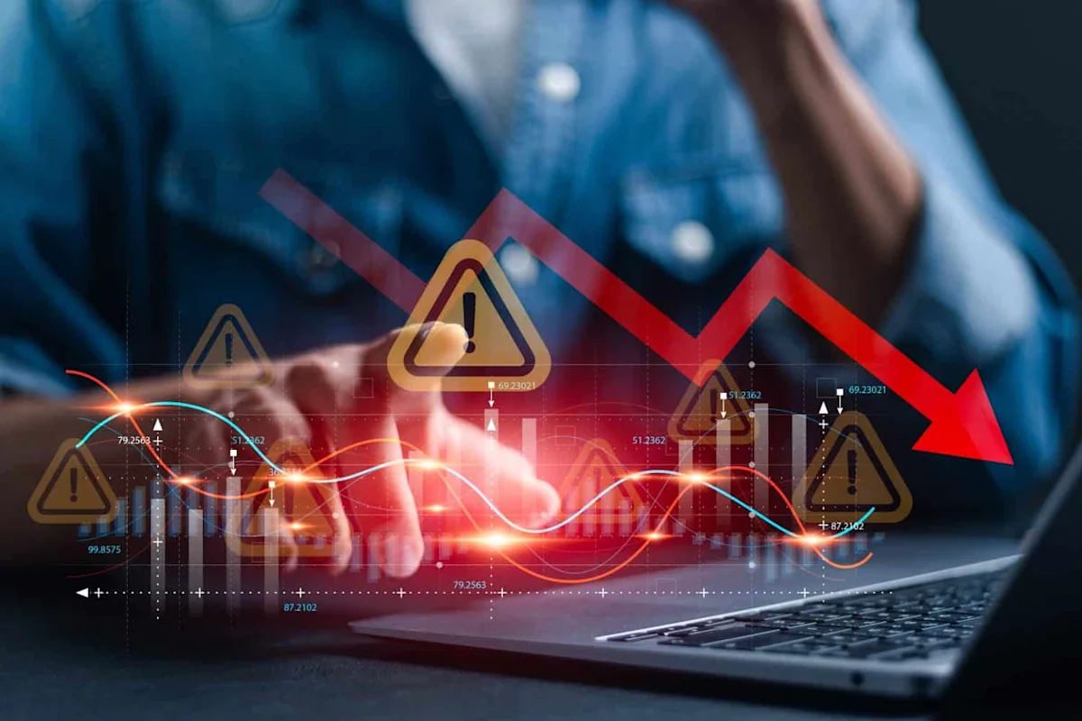 Person using a laptop with financial charts, warning symbols, and a sharply falling red arrow