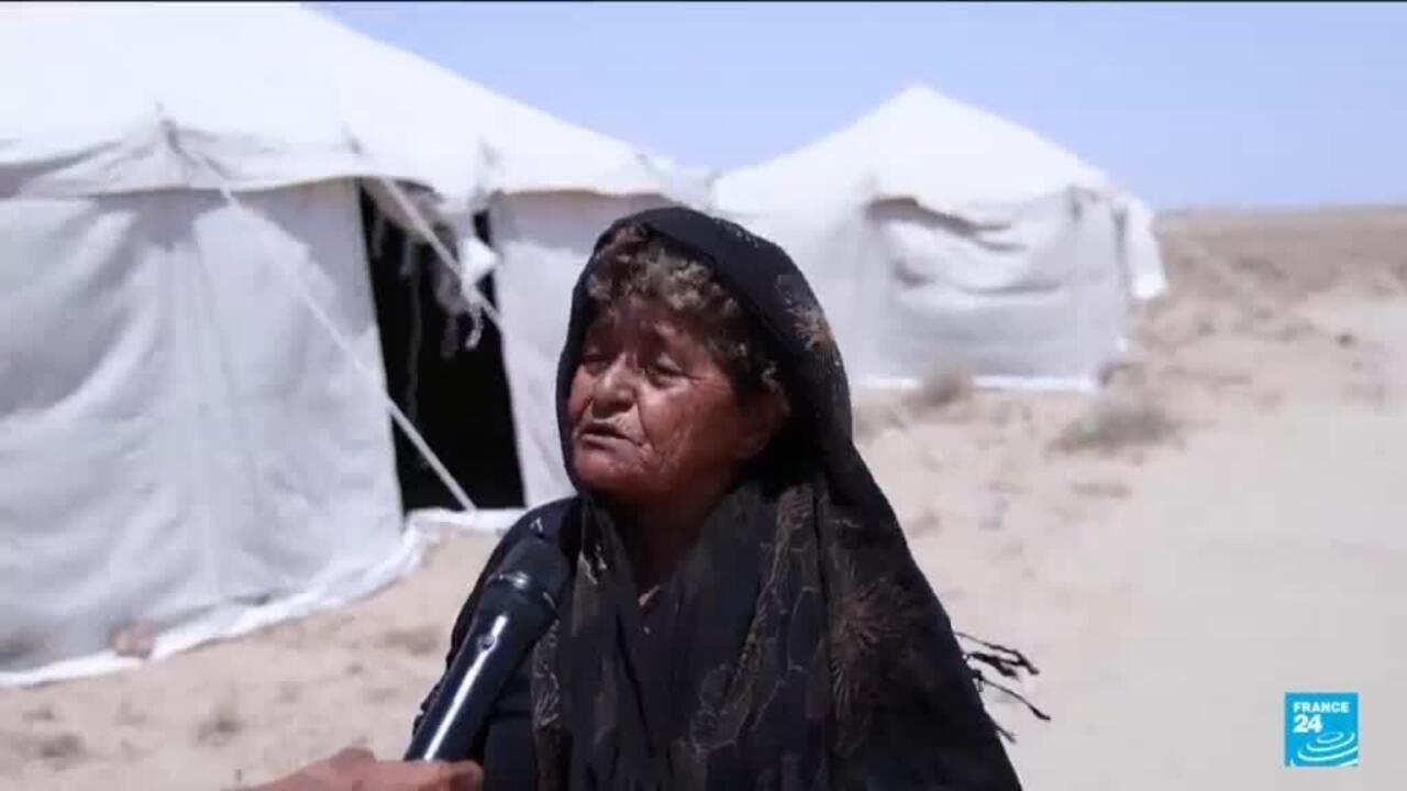 Woman in black headscarf speaks into a microphone beside white tents in a barren landscape.