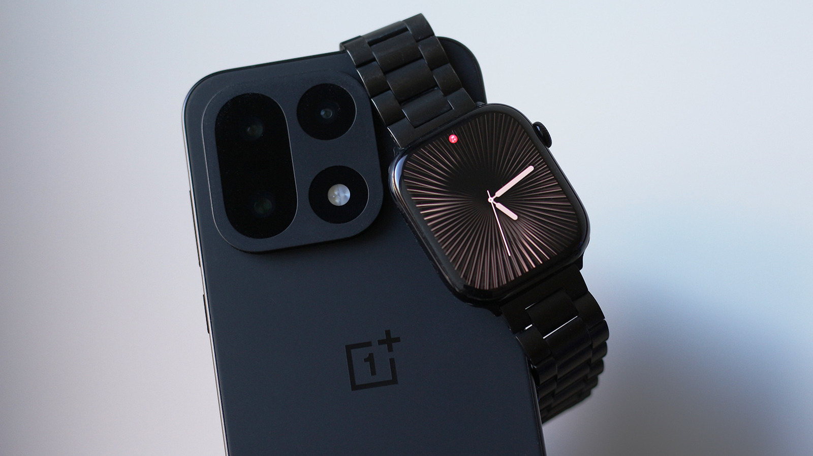 A black smartwatch with a metal band rests across the back of a black OnePlus smartphone.
