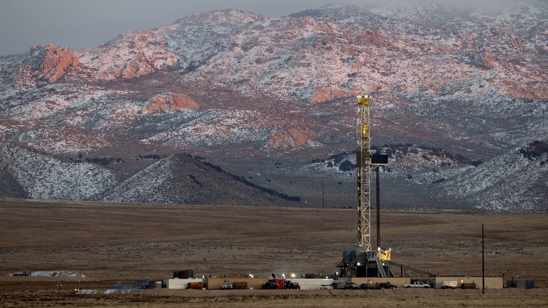 Drilling rig and support vehicles stand before snow-dusted red rock mountains and open farmland.