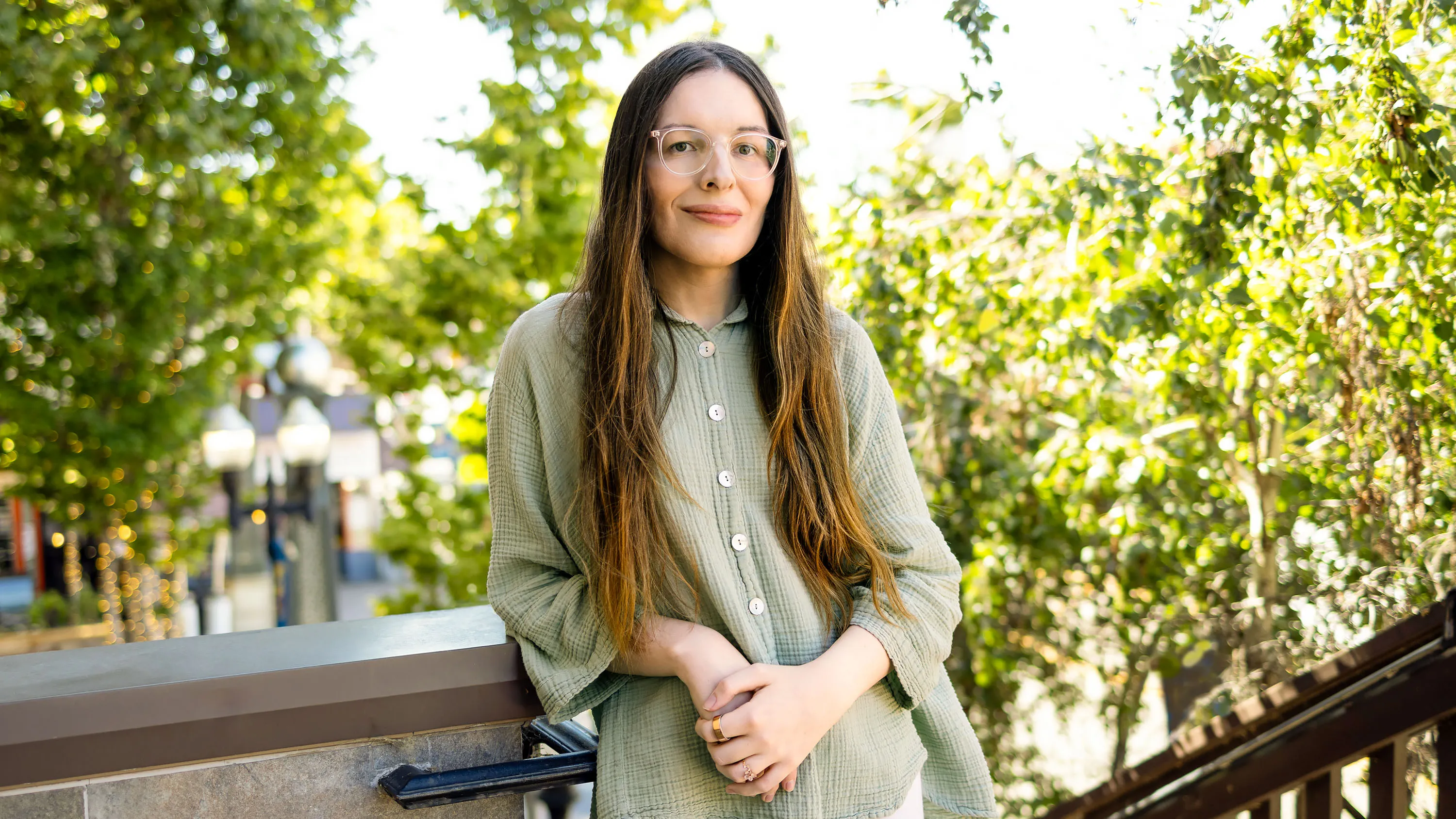 Woman wearing glasses and a sage-green shirt standing outdoors among leafy trees