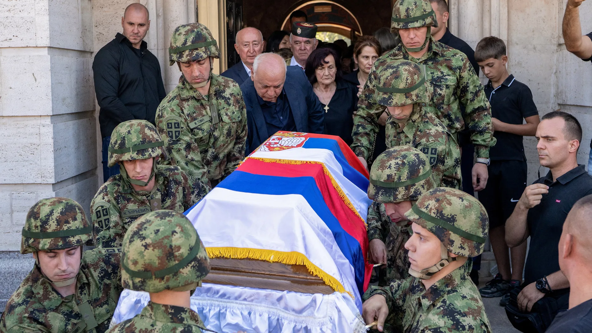 Soldiers carry a Serbian-flag-draped coffin from a building as mourners gather around them.