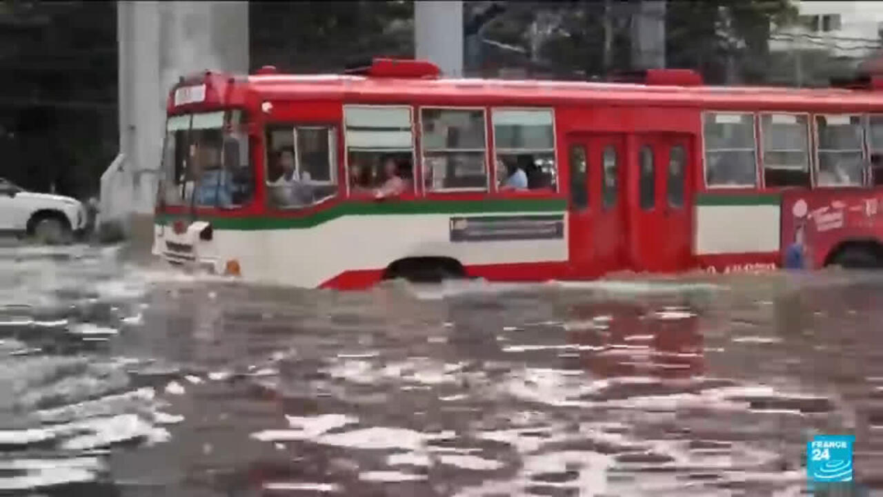 A red, white, and green city bus moves through deep, turbulent floodwater on a street.