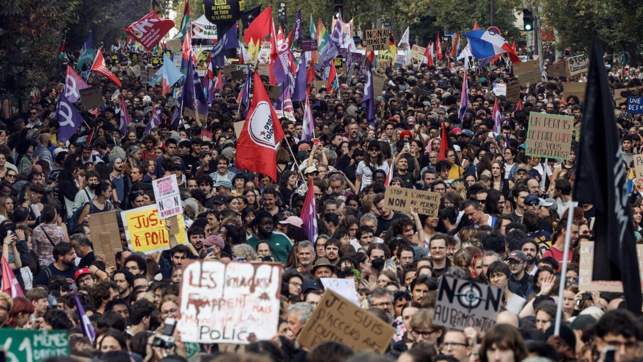 Large crowd of protesters carrying colorful flags and handwritten signs along a tree-lined street.