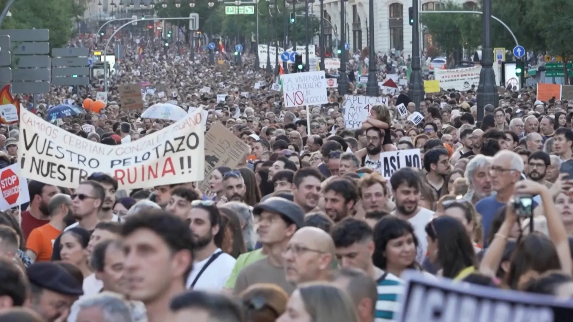 Dense street crowd holds protest signs, including messages about housing, amid Madrid-style buildings and traffic lights.