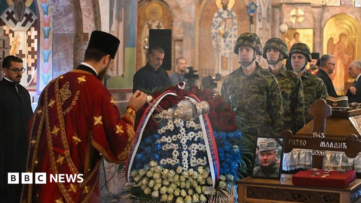 Clergyman conducts funeral ceremony as soldiers stand beside a flower-covered coffin in a church.