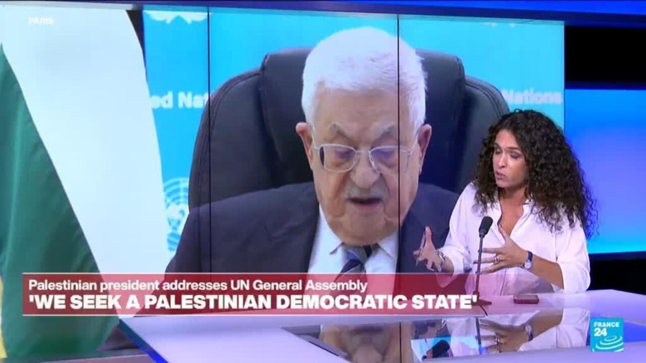 A woman speaks at a desk beside a large screen showing an older man addressing an assembly.